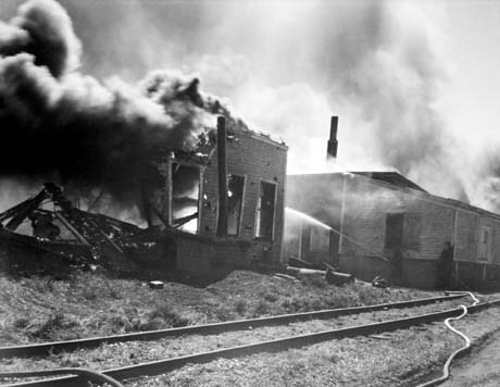 grayscale photograph: fireman spraying burning building with water, hoses and railway tracks in foreground