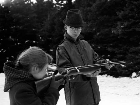 grayscale photograph: brother supports rifle while younger sister aims and shoots