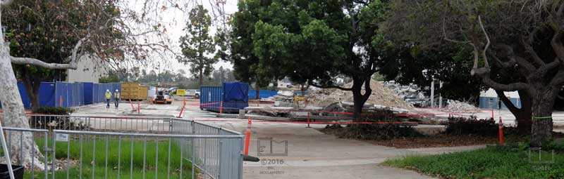 Walkway through grass and trees in foreground to workers, dumpsters and rubble piles in background