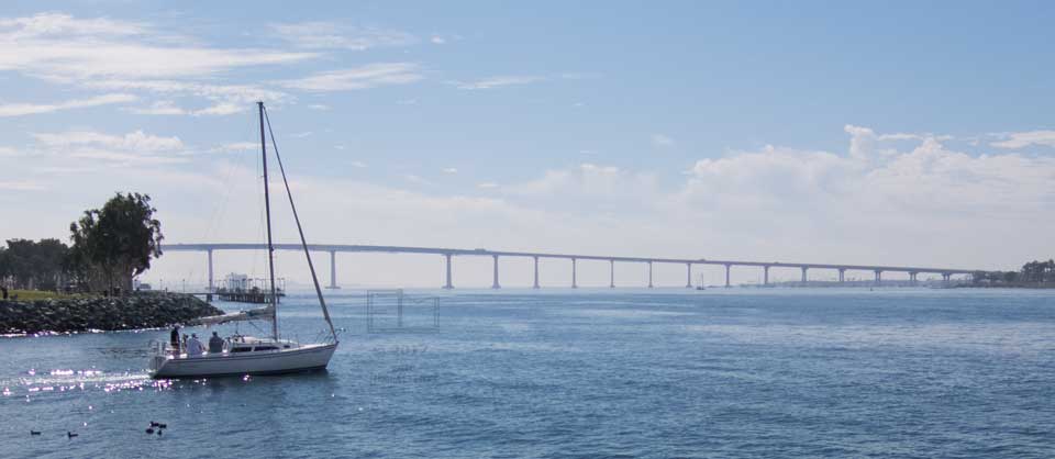 Small sailboat coming out of marina enterance withfull width of Coronado Bridge in background