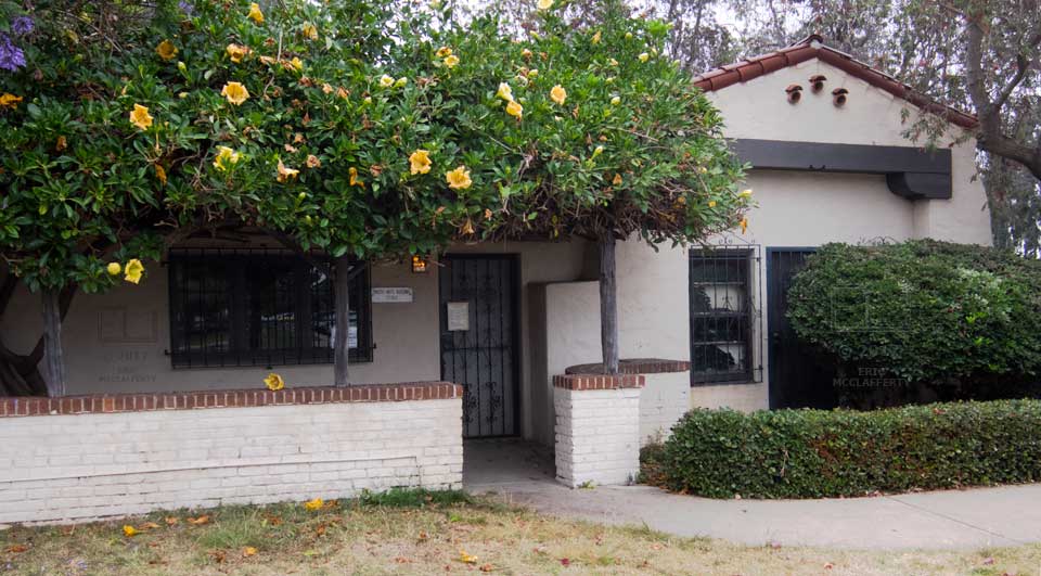 Small adobe building with large, flowered hedge overhanging entrance area