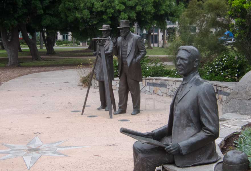 Three dark metal statues with surveying equipment, green park area in background