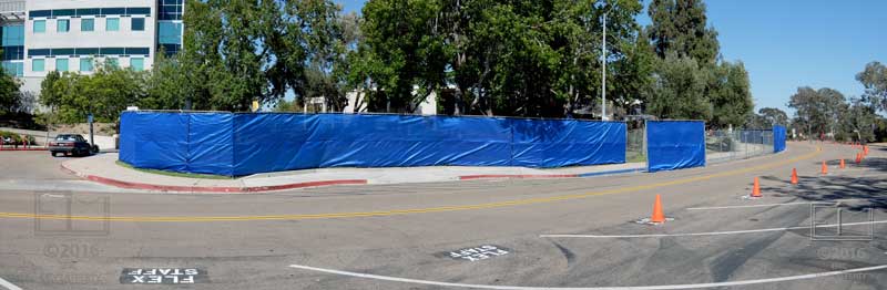 Wide view of blue fencing behind the old bookstore/cafateria buildings from across roadway