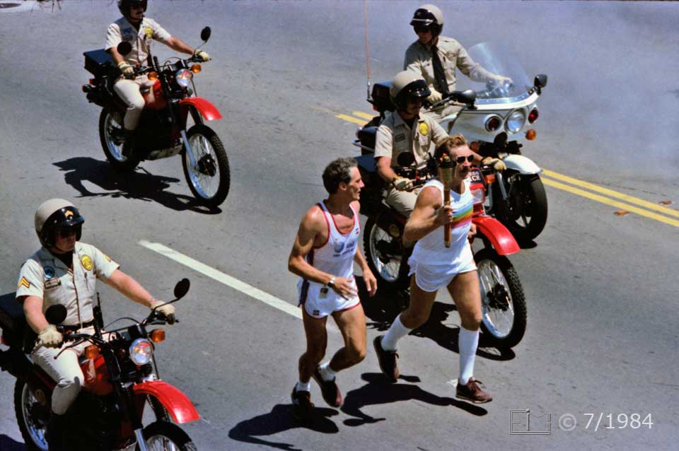 Color photo: Elevated view of running torch bearer, surouded by motercycle police  - Embedded text: 7/1984