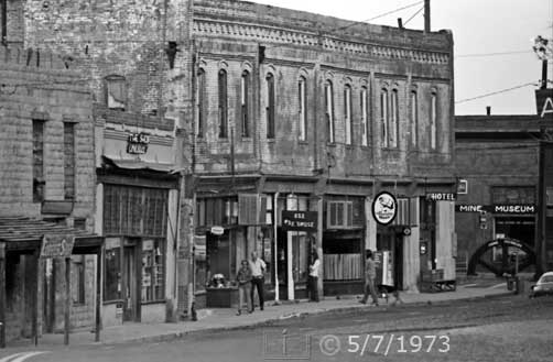 B/W photo: Oblique view of street with many storefronts and shops within old buildings - Embedded text: 5/7/1973