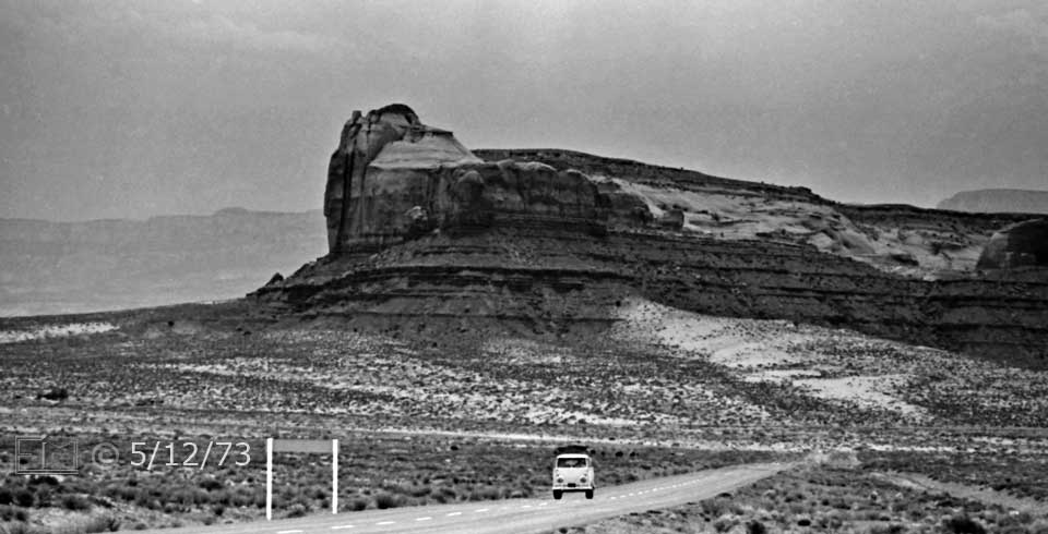 B/W photo: Mini-bus on a desert highway dwarfed by rock outcrop formation in background - Embedded text: 5/12/73