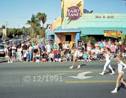 Color photo: Cheer leaders pass in front of curbside spectators and 'Daily Planet' bar - Embedded text: 12/1991