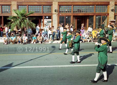 Color  photo: Wind instrument musicians, in colonial attire, in front of curbside spectators - Embedded text: 12/1991