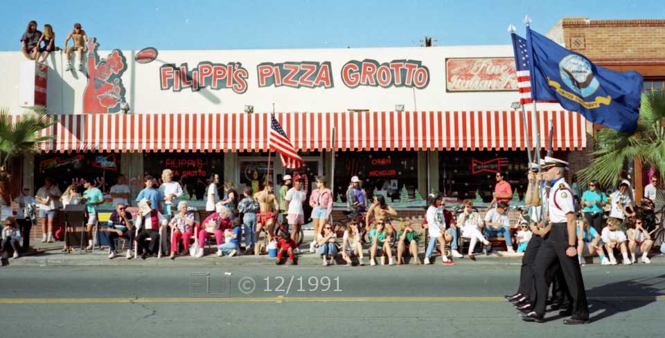 Color  photo: Male military marching, with flags, in front of Pizza Grotto being viewed from curbside and rooftop - Embedded text: 12/1991