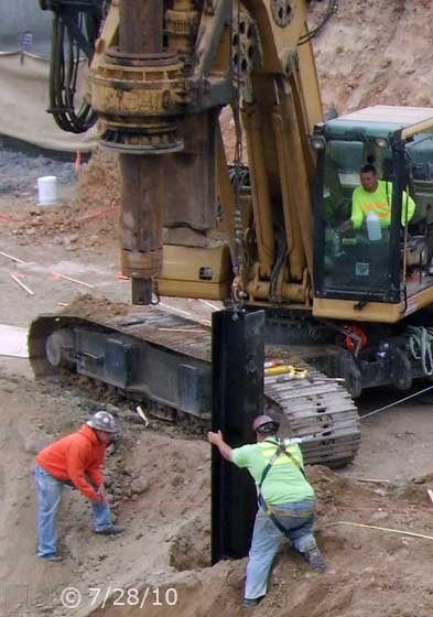 B/W photo: Workman and crane operator near completing loading of suspended I-beam into open posthole  - Embedded text: 7/28/10
