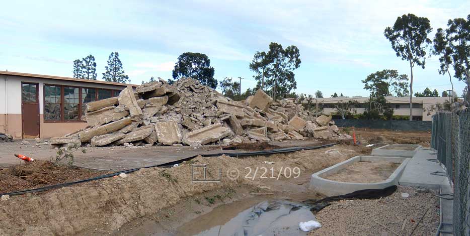 Color photo: Diagonal view of old building (west side, south end) with rubble pile and new buildings planters in front - Embedded text: 2/21/09