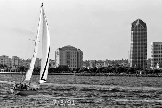 B/W photo: Stern view of boat under sail with San Diego skyline as background - Embedded text: 7/3/91