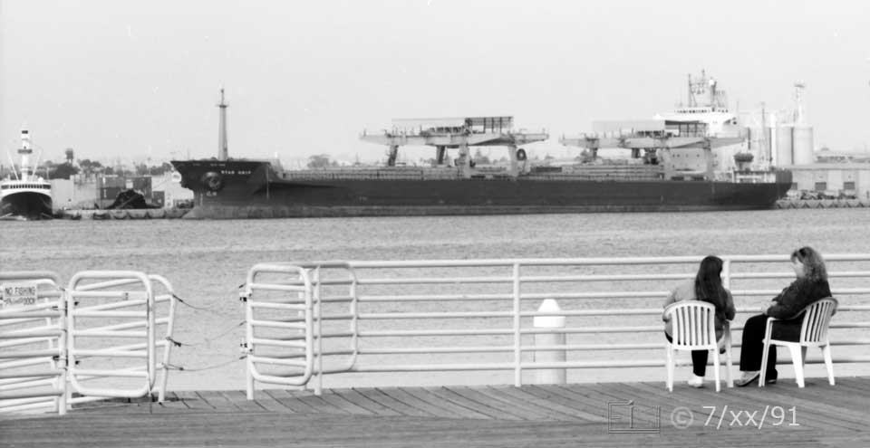 B/W photo: Two seated women conversing on a Coronado wharf witn tuna boat and large freighter as background on opposite side of bay - Embedded text: 7/xx/91